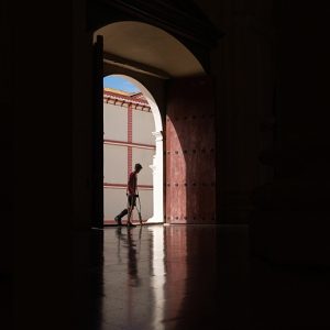 In a large, dark space, a young man using crutches passes a brightly lit doorway.