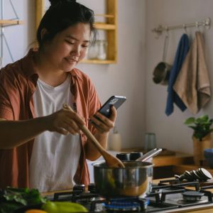 A young woman consults her phone as she stirs a saucepan