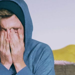Young white man in a gray hoodie rubs his face in distress.