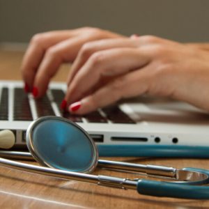 Close-up on a woman's hands as she types on a laptop. A stethoscope lies on the desk beside the computer.