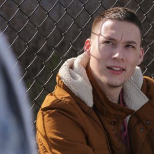 A young White man with short brown hair sits against a chainlink fence and looks up at a person who is out of focus and barely visible in the frame.