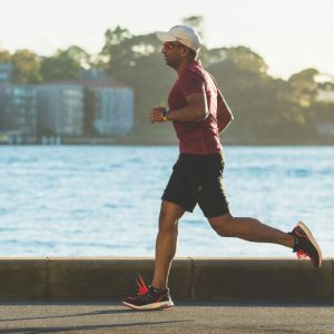 A man in a tee shirt, shorts, and a baseball cap jogs in front of a body of water.