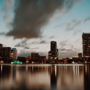 The skyline of downtown Orlando, Florida at dusk, with Lake Eola in the foreground and tall buildings behind it.