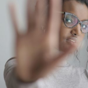 A young, Black woman in glasses holds. a hand up in a Stop" gesture, blocking the view of the camera