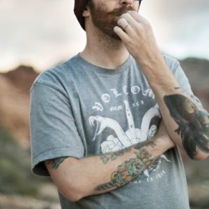 A man with tattoos on his arms and a Volcom t-shirt stands in front of a background of desert mountains. One hand is in front of his chin in a thoughtful pose.