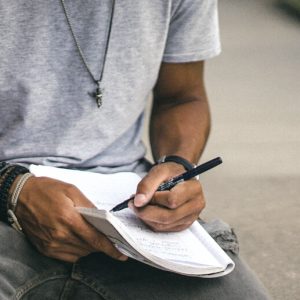 A man sits on a bench outdoors, writing in a notebook,