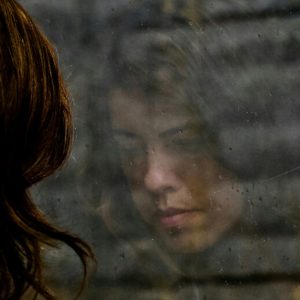 Reflection in the window of a sad-faced White woman with long hair. The view beyond her reflection is of a brick wall