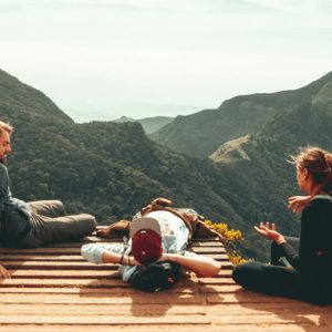 View from behind of three people sitting on a platform at the top of a mountain, with a view of other green peaks.
