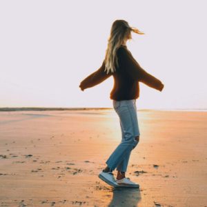 A woman in a sweater and jeans spins on the empty sands of a beach