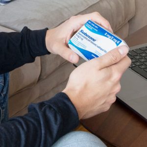 A man wearing a long-sleeved shirt and jeans looks at the Suboxone box held in his hands. He sits in front of an open laptop. Suboxone is sober.