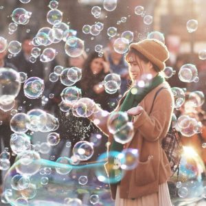 A woman wearing a knit hat, scarf, and jacket stands in a in a busy city space and laughs as she is surrounded by shimmering bubbles.