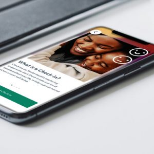 A smartphone sits face-up on a white table near a tablet and mug. The phone screen shows information about a Check-In in the Workit Health app