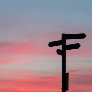 A street sign pointing in multiple directions is silhouetted against a pink and blue sunset