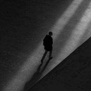 Black and white photo from above of a lone person crossing a wide, concrete space. The photo is moodily lit.
