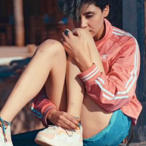 A young woman with short hair and an anxious expression sits on the floor with her knees pulled up to her chest.