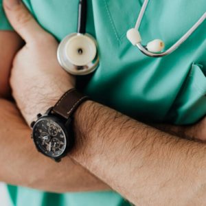 A doctor in mint green scrubs stands with his arms crossed. The camera does not show his face.