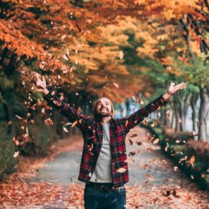 A man in a red plaid shirt stands with autumn trees behind him, throwing leaves in the air with a big smile on his face