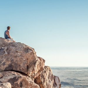 A person sits alone on a rocky outcropping, staring at the sea.