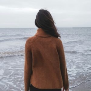 View of the back of a woman in a brown sweater as she stares out at the ocean.