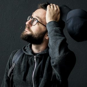 A man in a hoodie looks up questioningly, a baseball cap in his hand.