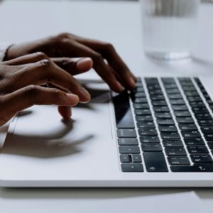 Dark-skinned hands type on a laptop on a clean, white desk.