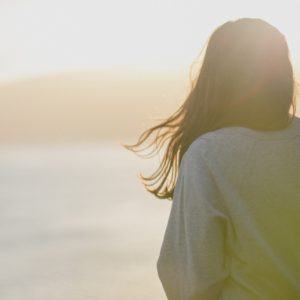 The back of a woman wearing a gray sweatshirt, sitting on a rocky shore and looking out at the sea. The sun is low and golden, as though it is very early morning or nearly sunset.