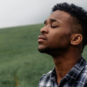 A young African American man stands in a grassy field, with his eyes closed and a pensive expression on his face.