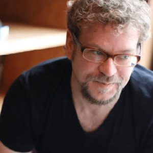 Author Daniel Maurer sits with a bookshelf behind him. He has curly hair and a goatee that are light brown. He wears glasses and a black t-shirt