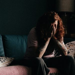 A person with long red hair sits on a sofa in a dark room with their head in their hands