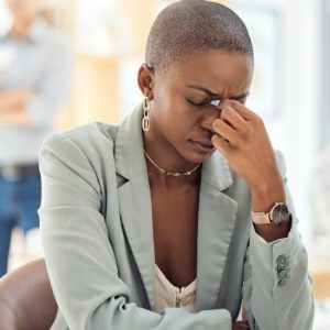 A young Black woman with close-cropped hair sits at a desk in a brightly lit office space. She pinches the bridge of her nose and looks distressed.