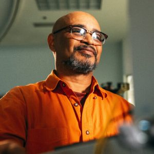 A man in an orange shirt and glasses stands at his work station facing a piece of electrical equipment.