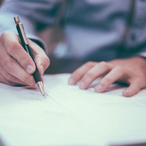 Closeup on the hands of a man in a long-sleeved, blue, button-up shirt as he writes on some papers with a ballpoint pen.