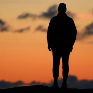 A man stands along on a hilltop, silhouetted by the red of the setting sun