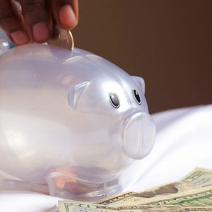 A clear plastic piggy bank sits on a white cloth. A brown-skinned hand is dropping a coin into the slot on the top of the bank, and there are $20 bills on the cloth in front of the bank.
