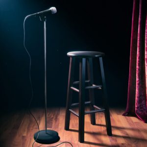 A black stool and microphone stand in a spotlight on an otherwise empty, dark stage.