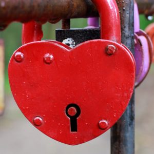 A red, heart-shaped padlock locked to a railing that also holds other padlocks.