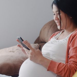 A young, East Asian woman who is clearly pregnant sits on a sofa and scrolls on her smartphone.