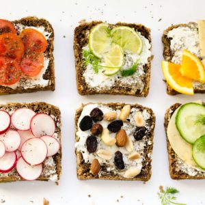 Six slices of whole whet bread arranged in a grid. Each is topped with goat cheese and additional ingredients, ranging from tomatoes to radishes to nuts.