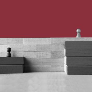 Wooden game pieces representing inequality. A dark piece stands on two stair-step blocks, facing the barrier of a high wooden wall. The light piece stands on top of the wall, with four stair-step blocks leading the way up.