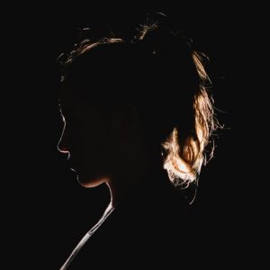 In a very dark space, a young woman is backlit so that her profile and ponytail are lit by a golden light, the only thing visible in the blackness.