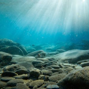 Rocky bottom to a body of water, with beams of light shining through the blue water.