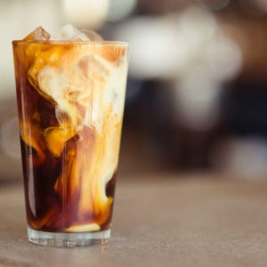 A glass tumbler of iced coffee with cream swirling through it.
