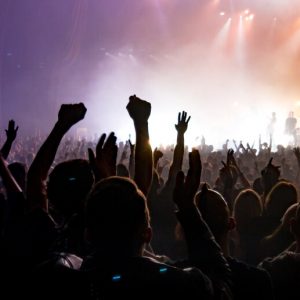 Rear view of the crowd at a concert with their hands in the air. The band is onstage in the distance, and the sky is hazy, with lights beaming through it.