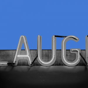 Neon sign reading "Laugh" against a blue background. Stand-up comedy in addiction recovery