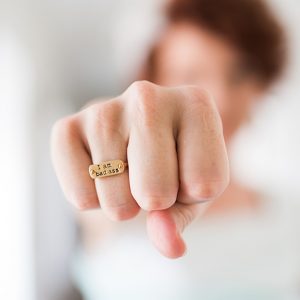 A woman in a white short holds her fist out determinedly toward the camera. She wears a metal ring stamped with the words "I am bad ass"