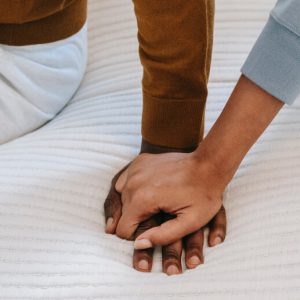 Focus on the hands of a couple sitting on a bed. The man's hand is pressed flat against the bed, and the woman's hand is on top of it, holding it.
