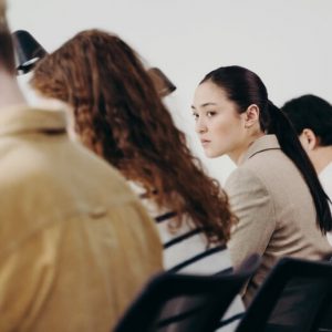 Office workers sitting in a row, seen from behind. One woman's face is in focus, as she looks toward her coworkers