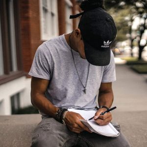 A man sits outdoors, writing in a notebook balanced on his leg