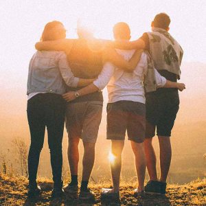 Four people with arms around one another stand atop a hill and look at the golden sunrise