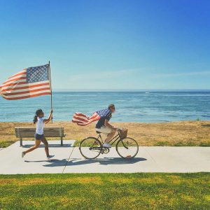 Beside the ocean, a man rides a bike with an American flag tied around his neck like a cape. Behind him, a woman runs with an American flag on a flagpole.